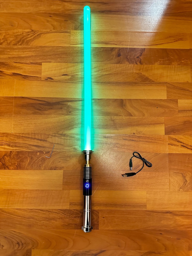 May include: A glowing green lightsaber with a metallic hilt featuring a black grip section and a blue illuminated power button. Included are a small L-shaped hex key and a black USB charging cable with a barrel plug connector for the device.