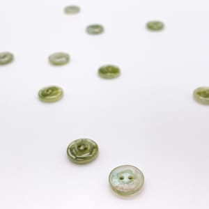 May include: A collection of round, iridescent buttons with two holes. The buttons have a pearlescent sheen with shades of green and white. The buttons are scattered on a white surface.