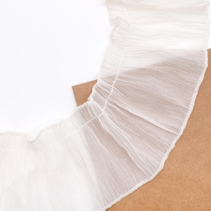 May include: A sheer white fabric trim featuring a delicate, crinkled texture and a gathered, ruffled edge. The material is lightweight and semi-transparent, showcasing a consistent pleated design along the length of the decorative textile strip.