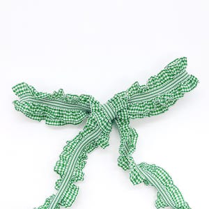 May include: A green and white gingham fabric strap featuring a ruffled edge and a central gathered section tied into a bow. The textile pattern consists of a small-scale checkered design throughout the entire length of the ruffled fabric.