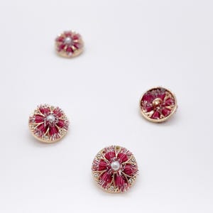 May include: Four circular buttons featuring a gold-toned metal base, a central white faux pearl, and radiating dark pink thread or fiber accents. The edges of the buttons are embellished with small, clear, reflective crystals arranged in a circular pattern.