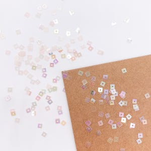 Könnte beinhalten: Eine Ansammlung kleiner, quadratischer, irisierender Pailletten, die auf einer weißen Oberfläche und einer Ecke einer braunen Kraftpapierkarte verstreut sind. Jede einzelne Paillette hat ein mittiges Loch und reflektiert Licht perlmuttartig.