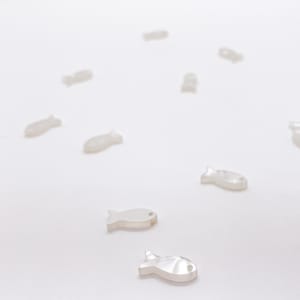 May include: A collection of small, white, iridescent fish-shaped charms scattered across a plain surface. Each charm features a smooth, pearlescent finish and a single small hole near the head, designed for use as a decorative component or bead.