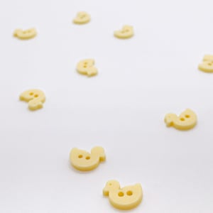 Peut inclure: Une collection de petits boutons jaune pâle en forme de canard. Chaque bouton comporte deux trous au centre pour la fixation. Les boutons en forme de canard sont dispersés dans le cadre, montrant leur surface lisse et leur silhouette.