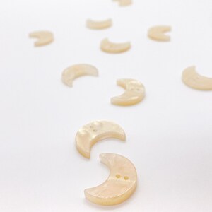 May include: A collection of small, crescent-shaped buttons. The buttons are a pale, pearlescent colour with two holes for sewing. The buttons are scattered on a white surface.