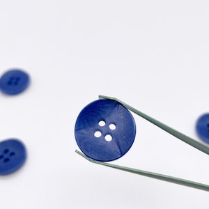 May include: A set of round, dark blue buttons, each featuring four holes in the center. One button is held by a pair of metal tweezers, while several other identical buttons are scattered around it on a plain, light-colored surface.