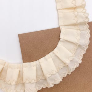 May include: Close-up of a cream-colored fabric trim with a ruffled design. The trim features a scalloped edge and is layered, creating a textured effect. The fabric is set against a white and brown background.