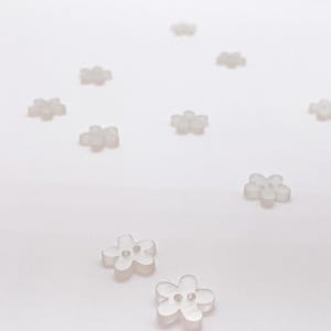 May include: A collection of small, translucent, clear plastic buttons shaped like five-petaled flowers. Each button features two small holes in the center for attachment. The buttons are scattered across the frame, showing a consistent floral design.
