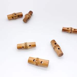 May include: Five cylindrical toggle buttons made of light brown bamboo with natural wood grain patterns. Each button features a central raised band and two small holes for threading, showcasing the organic texture and variations of the material.