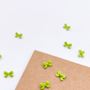 May include: A collection of small, bright lime green bow-shaped buttons. Several of these miniature bow buttons are scattered across the surface, while three are positioned on a piece of light brown textured cardstock paper.