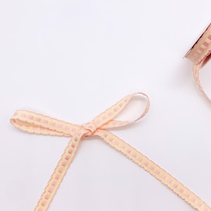 May include: A light yellow ribbon tied into a bow, featuring a repeating pattern of small pink squares along the center. The ribbon has a scalloped edge and is shown partially unspooled from a roll visible in the upper right corner of the frame.