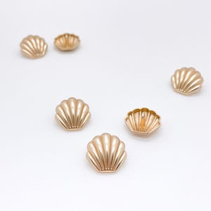 May include: A collection of five gold-toned metal buttons shaped like seashells. Each button features a fan-like design with raised vertical ridges and a central shank on the back for attachment, arranged in a scattered pattern.