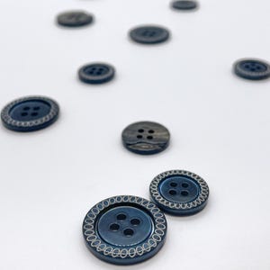 May include: Navy blue buttons with a silver decorative edge and four holes are scattered on a white surface. Some buttons are in focus, others blurred. The buttons vary in size and design. The buttons are suitable for clothing.