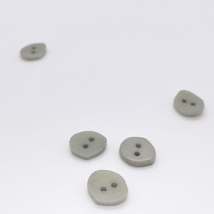 May include: Four small, smooth, light grey buttons with irregular, rounded shapes. Each button features two centred holes for attachment. The buttons are scattered across a plain, light-coloured surface, displaying a matte finish and simple design.