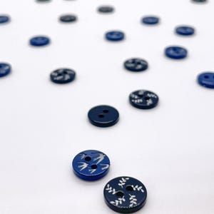 May include: A collection of navy blue and dark gray buttons scattered on a white surface. Some buttons feature white painted designs of birds or leaves. The buttons are round with two or four holes for sewing.