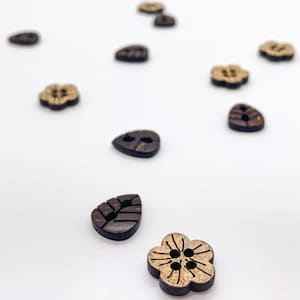 May include: A collection of wooden buttons in various shapes, including leaves and flowers, scattered on a white surface. The buttons are primarily dark brown with lighter brown accents, showcasing natural wood grain and textures. The buttons have two or four holes for sewing.