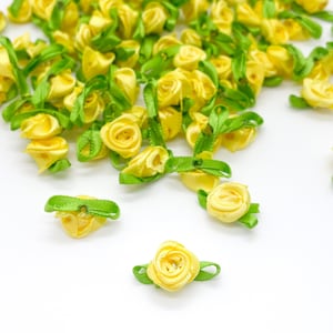 May include: A collection of small, yellow ribbon roses with green ribbon leaves. Each rose is crafted from folded satin-like fabric, featuring a tightly coiled centre and two small, pointed green leaves extending from the base of the flower.