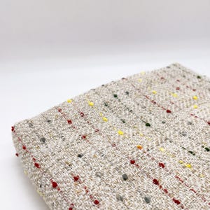 May include: A folded piece of beige fabric with a woven herringbone pattern. The textile features small, scattered flecks of red, yellow, green, and grey thread throughout the surface, creating a textured and multicolored appearance.