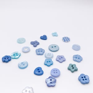 May include: A collection of assorted buttons in various shades of blue, including light blue, sky blue, and periwinkle. The buttons feature diverse shapes such as circles, flowers, hearts, and stars, with some displaying patterns like polka dots or bows.