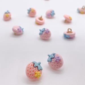 以下が含まれることがあります： ライトブルーとイエローのアクセントが付いた、ピンク色のイチゴ型のボタンのコレクション。ボタンはテクスチャ加工された表面で、白い背景に散らばっています。これらの装飾ボタンは、縫製やクラフトプロジェクトに最適です。
