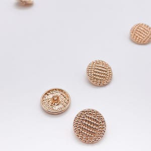 May include: A collection of five round, rose gold-coloured metal buttons. Each button features a textured, woven-like surface pattern on the front and a small metal loop shank on the back for attachment, displayed from various angles.