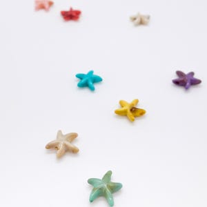 May include: A collection of small, five-pointed starfish-shaped beads scattered across a plain surface. The beads are rendered in various colours including pink, red, white, turquoise, purple, yellow, beige, and green with a smooth finish.