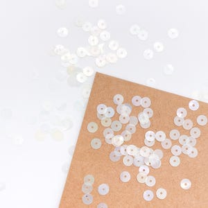 Puede incluir: Una colección de pequeñas lentejuelas blancas iridiscentes y circulares esparcidas sobre una superficie blanca lisa y un trozo rectangular de papel kraft marrón claro. Cada lentejuela tiene un orificio central y un acabado translúcido.