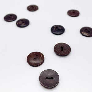 May include: A collection of ten round, dark brown buttons with a textured, mottled surface. Each button features two small holes in the center for attachment. The buttons are scattered across the frame, showing variations in their natural pattern.