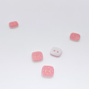 May include: Four square buttons with rounded corners, featuring two central holes. Three buttons are pink with a white polka dot pattern, and one button is solid white. The buttons are arranged in a scattered formation on a plain surface.
