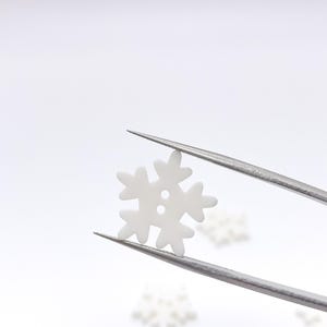 May include: A small, white, snowflake-shaped button with two holes in the center. The button is held by a pair of metal tweezers, showcasing its flat, smooth surface and six-pointed design, with additional identical buttons visible below.