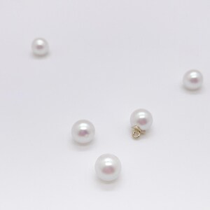 May include: Five smooth, spherical white pearls arranged on a plain surface. Four of the pearls are loose, while one pearl features a small, gold-toned metal loop attached to its base, designed for use as a jewelry component or charm.