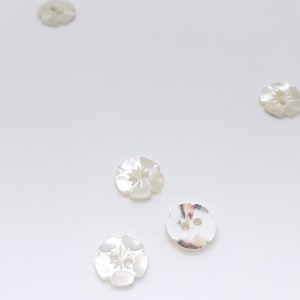 May include: A collection of five small, round buttons with a pearlescent, off-white finish. Four of the buttons feature a carved, five-petaled flower design, while one button is smooth with two holes and subtle, multi-colored marbled patterns.