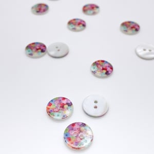 Op de afbeelding: Een verzameling ronde knopen met twee gaten verspreid over het beeld. Elke knoop heeft een gladde, witte, iriserende afwerking aan de ene kant en een levendig, veelkleurig bloemenpatroon aan de achterkant, met tinten roze, blauw en groen.