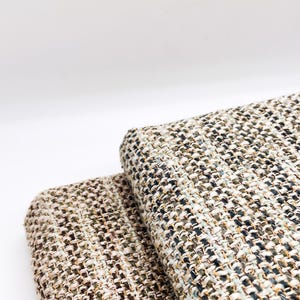 May include: Two folded pieces of fabric featuring a thick, textured weave. The material displays a complex blend of neutral tones, including beige, cream, dark brown, black, and subtle hints of sage green, creating a variegated, multi-colored appearance.