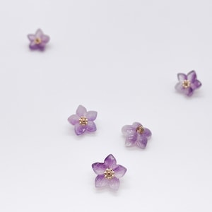 May include: Five small, five-petaled flower-shaped beads made of translucent purple amethyst stone. Each flower features a central gold-toned metal component with a textured, circular design at its core, creating a delicate floral appearance.