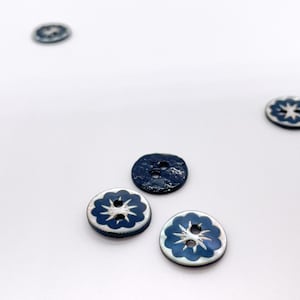 Puede incluir: Varios botones florales azul marino y blancos sobre una superficie blanca. Los botones tienen un borde festoneado y dos agujeros para coser. Los botones presentan texturas y acabados variados.