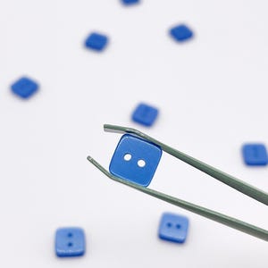 Può includere: Un set di piccoli bottoni quadrati blu con due fori al centro. Un bottone è tenuto da una pinzetta metallica, mentre diversi altri bottoni blu identici sono sparsi attorno su una superficie bianca uniforme, caratterizzati da una finitura liscia.