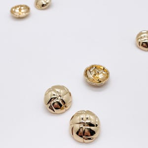 May include: A collection of five round, gold-coloured buttons with a quilted, cross-hatched surface texture. Each button features a polished, reflective metallic finish and a shank-style attachment point on the underside for sewing onto garments.