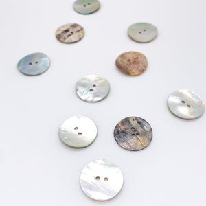 May include: A collection of ten round buttons made from iridescent shell material. Each button features two small holes in the center. The buttons display various natural patterns and colors, including shades of white, cream, gray, and brown.