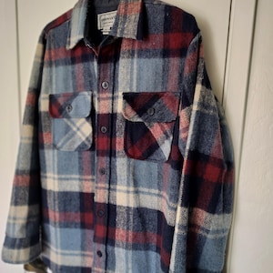 May include: A long-sleeve button-up shirt featuring a plaid pattern in shades of navy blue, red, and cream. The shirt includes a pointed collar, two chest pockets with button flaps, and a central button placket. A label reads Oshman's.