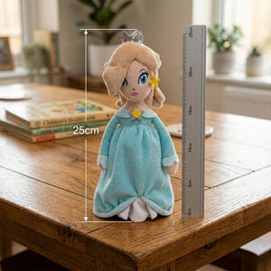 May include: A plush doll of a character with blonde hair, blue eyes, and a crown. The doll is wearing a light blue dress and is approximately 10 inches tall. A ruler is placed next to the doll to measure its height.