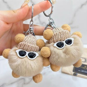Puede incluir: Dos llaveros de felpa beige, redondos y esponjosos, cada uno con un pequeño gorro de punto color canela y gafas de sol con montura blanca y lentes negras. Las figuras de felpa tienen orejas pequeñas y redondas y anillas de metal plateado.
