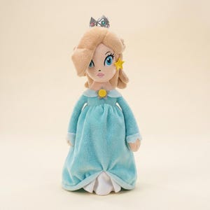May include: A plush doll of a character with blonde hair, blue eyes, and a crown. The doll wears a light blue dress with long sleeves and a white underskirt. A yellow star-shaped accessory is on the doll's hair.