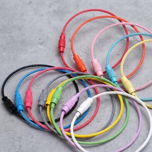 May include: A collection of circular wire cable keychains featuring flexible, plastic-coated steel loops in various colors including red, pink, blue, yellow, green, purple, white, gray, and black, each secured with a threaded metal barrel connector.