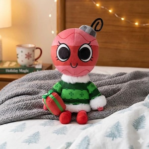 K&ouml;nnte beinhalten: Ein Pl&uuml;sch-Weihnachtsschmuck-Charakter mit einem rosa und rot gestreiften Gesicht, gro&szlig;en schwarzen Augen und einem l&auml;chelnden Mund. Er tr&auml;gt einen gr&uuml;nen Pullover mit wei&szlig;em Besatz und h&auml;lt eine rote Geschenkbox. Die Figur liegt auf einem Bett mit einer grauen Decke.