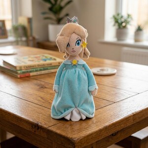 May include: A plush doll of a character with blonde hair, a silver crown, and a light blue dress. The doll has a yellow star-shaped earring and a yellow brooch. The doll is standing on a wooden table.