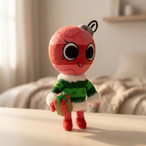 K&ouml;nnte beinhalten: Ein Pl&uuml;sch-Weihnachtsschmuck-Charakter mit einem roten und rosa Gesicht, gro&szlig;en schwarzen Augen und einem l&auml;chelnden Mund. Er tr&auml;gt einen gr&uuml;n-wei&szlig;en Pullover und h&auml;lt ein kleines Geschenk. Der Schmuck hat rote F&uuml;&szlig;e und eine silberne Oberseite.