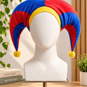 Pode incluir: Um chapéu de bobo da corte com um esquema de cores vibrantes de painéis de veludo alternados em vermelho e azul. O chapéu inclui uma faixa amarela brilhante na base e duas pontas longas e curvas que caem nas laterais, com pompons amarelos.