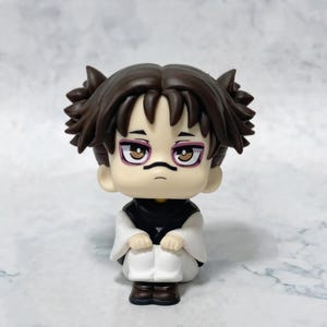 Könnte beinhalten: Eine kleine Figur im Chibi-Stil, die einen Charakter mit dunkelbraunem Haar in zwei seitlichen Knoten, heller Haut und braunen Augen darstellt. Die Figur trägt ein schwarzes Oberteil mit weißen Ärmeln und eine weiße Hose und sitzt mit neutralem Blick.