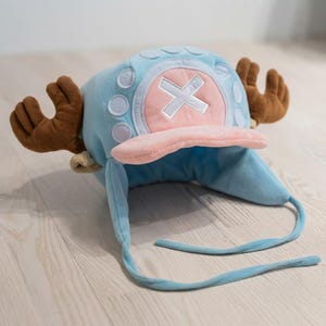 May include: A light blue plush hat featuring brown reindeer antlers on the sides, a pink brim, and a white cross emblem on the front. The hat includes ear flaps with long light blue ties hanging down from the sides and circular accents.
